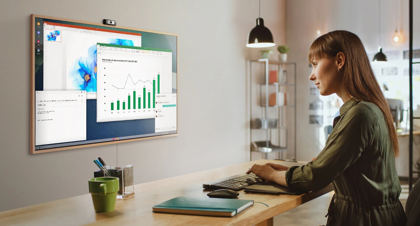 A woman is using her office PC to do work on her TV screen at home. On the screen of The Frame are various office productivity windows.